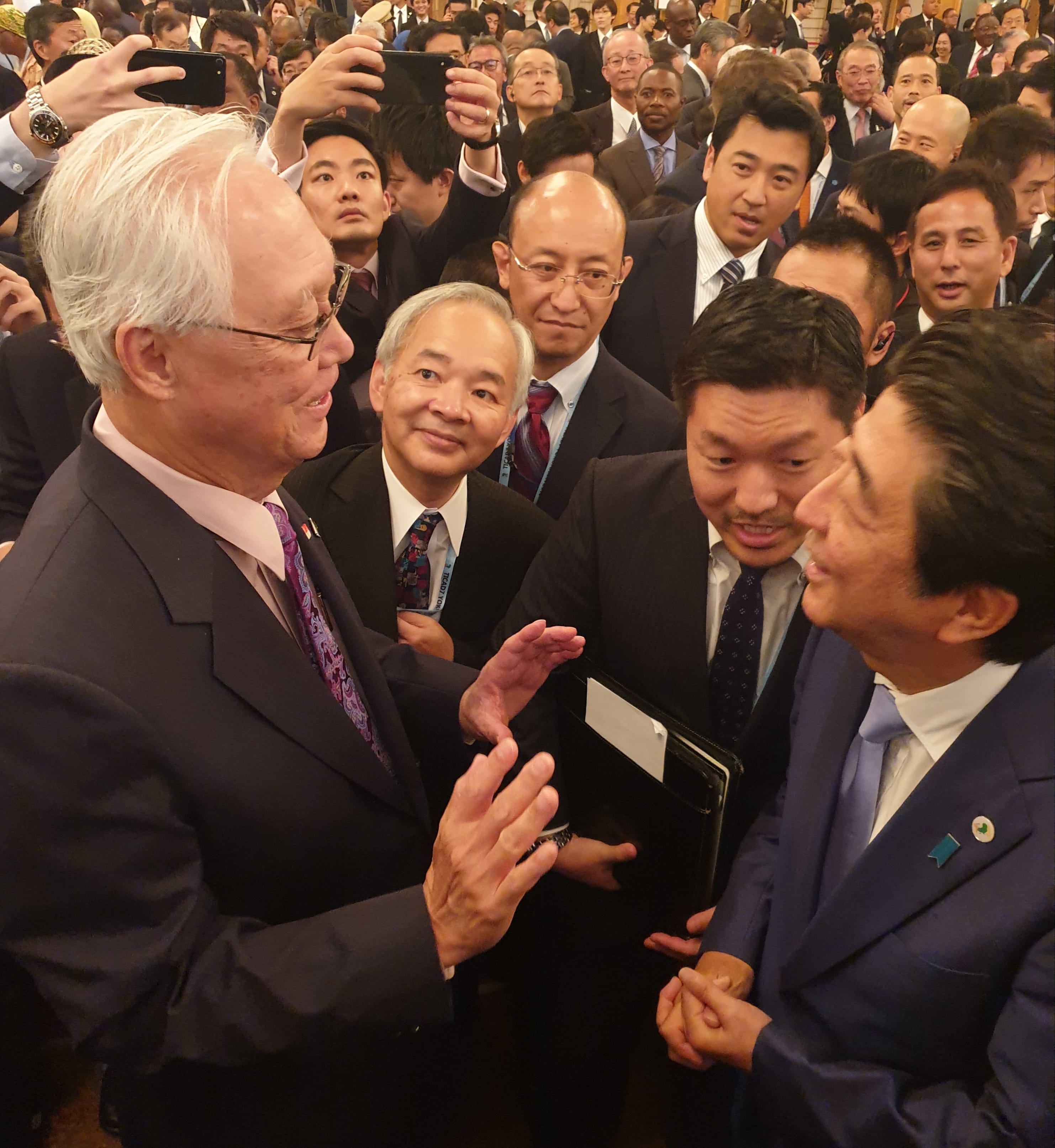 Shinzo Abe in a suit, surrounded by a crowd, shakes hands with a man with white hair.
